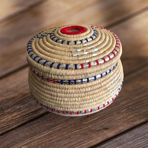 May include: A round woven basket with a lid. The basket is made of natural fibres and has a decorative trim of silver and red ribbon. The lid has a red centre.