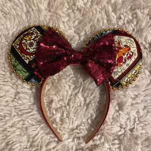 Könnte beinhalten: Ein Paar Ohren im Maus-Design mit einer bordeauxfarbenen Pailletten-Schleife und einem roten Stirnband. Die Ohren sind mit farbenfrohen Buntglasmustern verziert und mit goldenen Perlen umrandet. Sie liegen auf einem weißen, flauschigen Hintergrund.