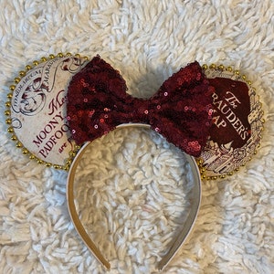 Könnte beinhalten: Ein Stirnband mit einer burgunderfarbenen Pailletten-Schleife und zwei runden Ohren. Die Ohren haben ein Kartendesign mit goldenem Rand und Text. Das Stirnband ist weiß und goldfarben. Die Schleife ist mittig auf dem Stirnband angebracht.