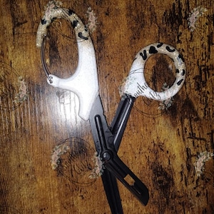 Może przedstawiać: Czarno-białe nożyczki medyczne z ozdobnym uchwytem z nadrukiem w cętki i brokatowymi akcentami. Nożyczki mają zakrzywione ostrze i zabezpieczoną końcówkę. Na uchwycie znajduje się również napis "Southern Glitz".