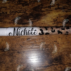 Könnte beinhalten: Ein weißer und goldener Glitzer-Stift mit einem schwarzen Leopardenmuster. Der Stift ist mit dem Namen "Nichole" in schwarzer kursivschrift personalisiert.
