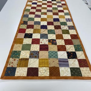 May include: A rectangular patchwork quilt with a checkerboard pattern. The quilt features squares of various fabrics in shades of brown, red, green, and cream, with a brown border. The quilt is likely used as a table runner or decorative piece.