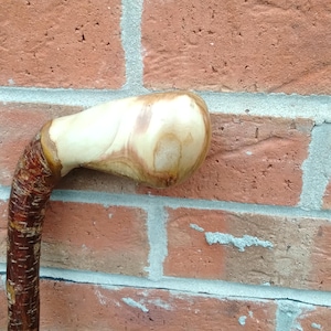 May include: A wooden walking stick with a light-coloured, rounded handle and a dark brown, textured shaft. The handle has a natural wood grain pattern. The walking stick is leaning against a red brick wall.