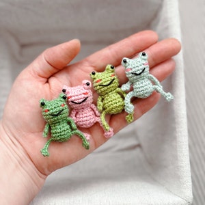 May include: Four crocheted frog figurines in a white box. The frogs are green, pink, and light green. They are sitting on a hand.