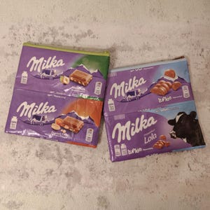 Peut inclure: Deux pochettes zippées violettes Milka. L'une avec une fermeture éclair verte et une barre chocolatée aux noisettes. L'autre avec une fermeture éclair bleue et une barre chocolatée avec une vache.