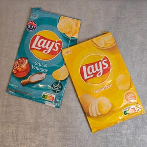 Könnte beinhalten: Zwei Tüten Lay's Kartoffelchips. Eine ist türkis mit dem Text "Salt & Vinegar" und einer Grafik von Salzstreuer und Essigflasche. Die andere ist gelb mit dem Text "Gesalzen" und einer Kartoffelgrafik. Beide haben das Lay's-Logo.