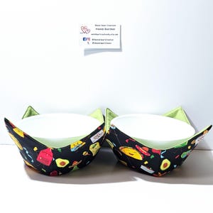 May include: Two white ceramic bowls nestled in black fabric bowl cozies. The cozies feature a colorful pattern of tacos, avocados, and other food items. The interior of the cozies is a light green color.