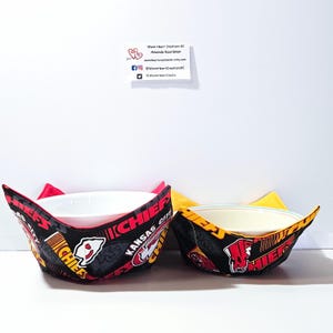 May include: Two fabric bowl cozies in black, red, and yellow with the Kansas City Chiefs logo and text. The larger cozy is red and the smaller cozy is yellow.