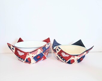 May include: Two fabric bowl cozies in red, white, and blue patriotic colors with a star and heart pattern. The cozies are holding white bowls.
