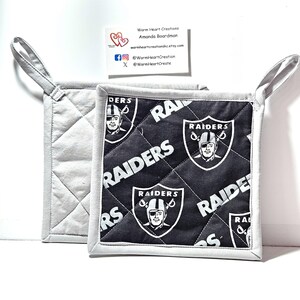 May include: Two square quilted pot holders featuring a black fabric with a repeating white Las Vegas Raiders logo and the word RAIDERS. The pot holders have light gray trim and a matching gray quilted back with a fabric loop for hanging.