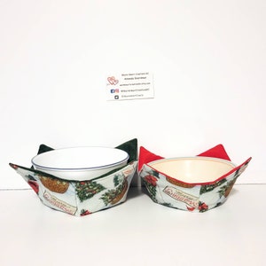 May include: Two Christmas-themed bowl cozies, one with a green trim and one with a red trim. Both cozies feature a white background with a repeating pattern of green pine branches, red berries, and a red and white "Merry Christmas" banner. The cozies are holding white bowls.