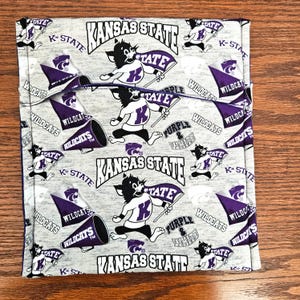May include: A light gray fabric featuring a repeating pattern of Kansas State Wildcats logos, mascots, purple pennants, and megaphones. The text includes Kansas State, Wildcats, and Purple & White. The design is printed in purple, black, and white.