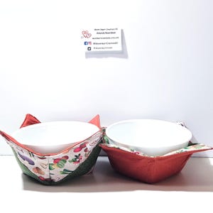 Puede incluir: Dos cuencos blancos con calientaplatos de tela roja y verde. Los calientaplatos tienen un patrón floral con verduras. Los calientaplatos están diseñados para mantener los cuencos calientes o fríos.
