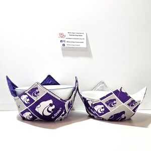 May include: Two purple and gray fabric bowl cozies with the K-State Wildcats logo. The cozies are designed to hold a bowl and keep it warm.