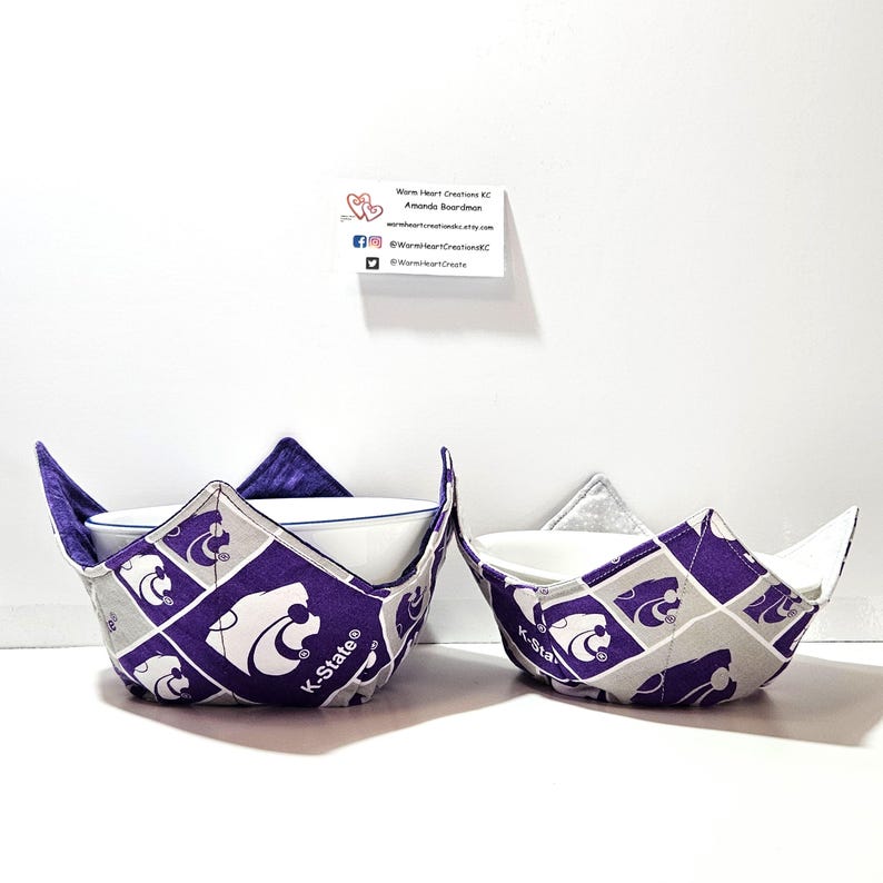May include: Two purple and gray fabric bowl cozies with the K-State Wildcats logo. The cozies are designed to hold a bowl and keep it warm.