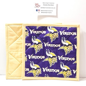 Puede incluir: Dos salvamanteles cuadrados acolchados. Uno es de color amarillo claro liso, el otro presenta un fondo morado con un patrón repetido de una cabeza vikinga y la palabra "VIKINGS" en blanco. Los bordes están ribeteados en amarillo claro. El texto en una tarjeta blanca dice "Warm Heart Creations".