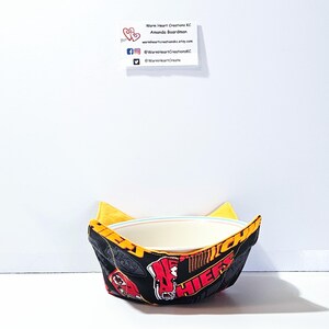 May include: A yellow and black bowl cozy with a red and white Chiefs logo. The cozy is holding a white bowl.