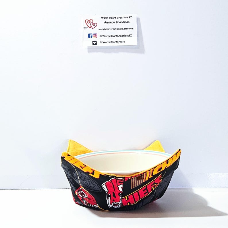 May include: A yellow and black bowl cozy with a red and white Chiefs logo. The cozy is holding a white bowl.