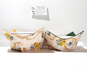 May include: Two fabric bowl cozies with a floral and bee design. The cozies are folded and displayed on a white surface. The cozies are made of a light pink fabric with yellow flowers, green leaves, and black and yellow bees. The text "Warm Heart Creations KC" is visible on a business card in the background.