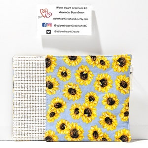 May include: Two blue and white fabric pot holders. One side has a white background with a grid pattern. The other side has a blue background with a yellow sunflower pattern. The text "Warm Heart Creations KC" is on a business card in the background.