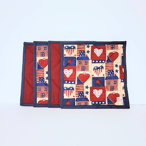 May include: Set of three patriotic placemats. Each placemat features a repeating pattern of red hearts, blue stars, and houses on a cream background. The edges are trimmed with a navy blue border and a red backing.