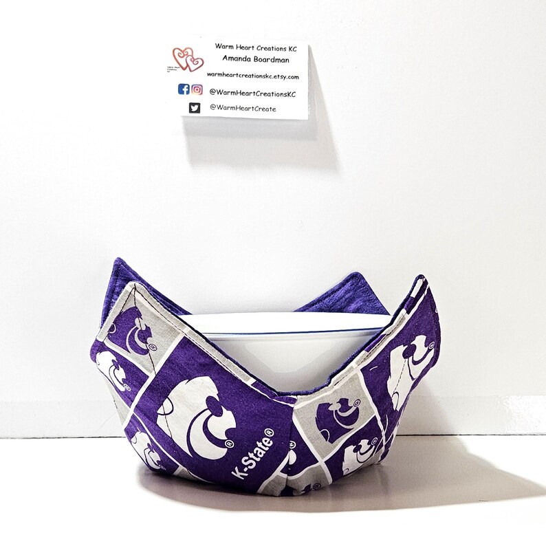 May include: Purple and gray bowl cozy with a K-State logo. The cozy is made of fabric with a repeating pattern of the K-State Wildcat mascot.