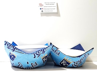 May include: Two blue and white bowl cozies with a Kansas City Royals logo pattern. The cozies are made of fabric and have a quilted design. The cozies are sitting on a white surface.