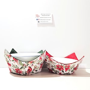 May include: Two red and green Christmas-themed bowl cozies with white bowls inside. The cozies feature a red cardinal, holly, and poinsettia pattern.