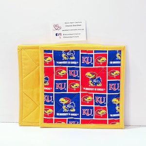 May include: Two square quilted pot holders. One is yellow, the other has a red, blue, and yellow pattern with the text "KU" and "The University of Kansas" and a Jayhawk logo. The pot holders are handmade.