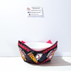 May include: A black and red fabric bowl cozy with the Kansas City Chiefs logo and the words "Kansas City Chiefs" repeated around the bowl cozy. The bowl cozy is holding a white ceramic bowl.
