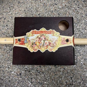 May include: A cigar box guitar with a dark brown wooden body, a light-colored wooden neck, and a decorative label featuring the text "MARIA MANCINI" and a classical scene. The guitar has a round sound hole.