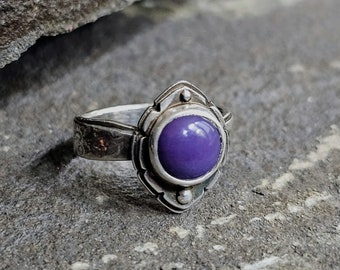 May include: A silver ring featuring a purple gemstone set within an intricate, ornate design.