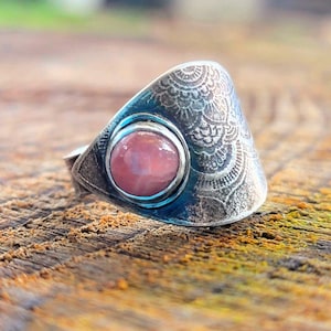 May include: A silver ring featuring a pink gemstone set in a bezel setting. The ring band is textured with a floral design.