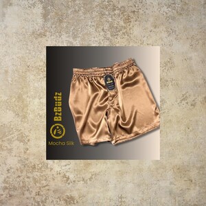 May include: A pair of brown satin boxer shorts with a black elastic waistband and a small black tag that says "BZBuddz". The tag is located on the right side of the waistband. The shorts are made of a soft, silky material and have a relaxed fit.