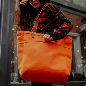 K&ouml;nnte beinhalten: Eine gro&szlig;e orangefarbene Canvas-Tragetasche mit braunen Ledergriffen. Die Tasche hat eine gro&szlig;e Tasche auf der Vorderseite und ist aus strapazierf&auml;higem Material gefertigt.