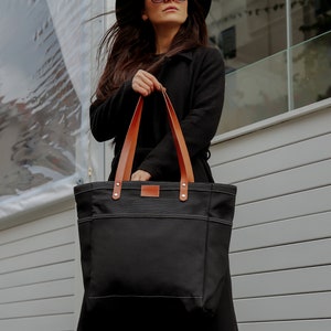 K&ouml;nnte beinhalten: Eine schwarze Canvas-Tragetasche mit braunen Lederriemen. Die Tasche hat eine gro&szlig;e Tasche auf der Vorderseite und wird von einer Person in einem schwarzen Mantel gehalten.