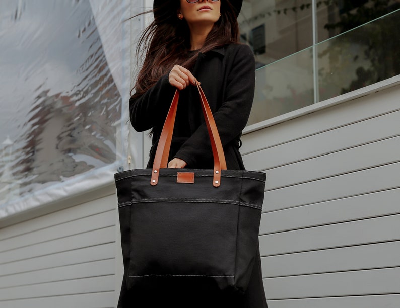 K&ouml;nnte beinhalten: Eine schwarze Canvas-Tragetasche mit braunen Lederriemen. Die Tasche hat eine gro&szlig;e Tasche auf der Vorderseite und wird von einer Person in einem schwarzen Mantel gehalten.