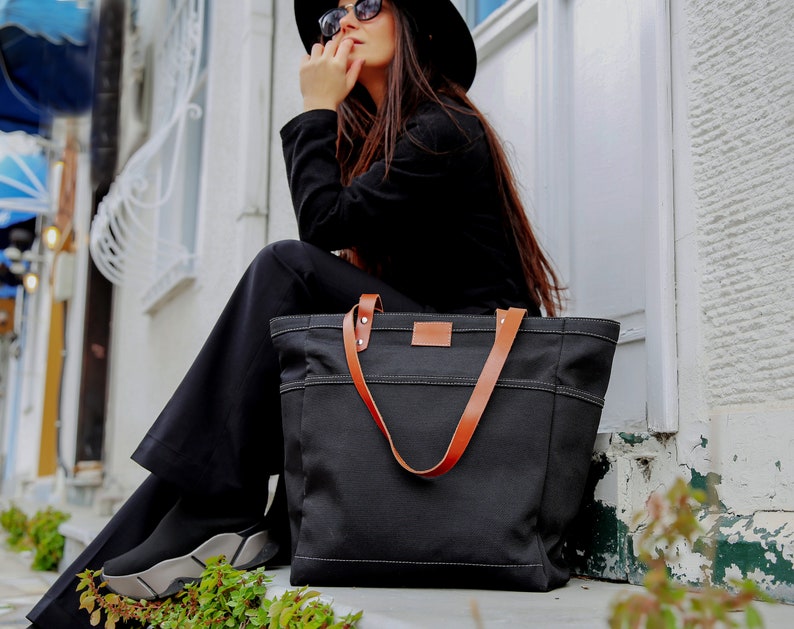 K&ouml;nnte beinhalten: Eine schwarze Canvas-Tragetasche mit braunen Ledergriffen. Die Tasche hat einen Rei&szlig;verschluss und ein gro&szlig;es Hauptfach.