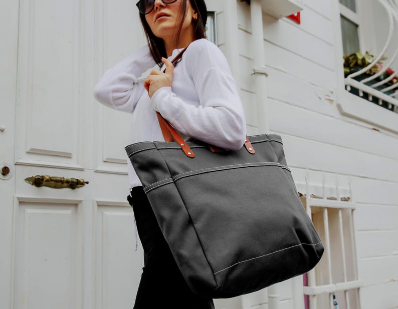 K&ouml;nnte beinhalten: Eine graue Canvas-Tragetasche mit braunen Ledergriffen. Die Tasche hat eine gro&szlig;e Tasche auf der Vorderseite und einen Rei&szlig;verschluss.
