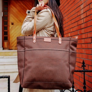 May include: A brown canvas tote bag with leather straps and a brown leather patch on the front. The bag is being held by a person.