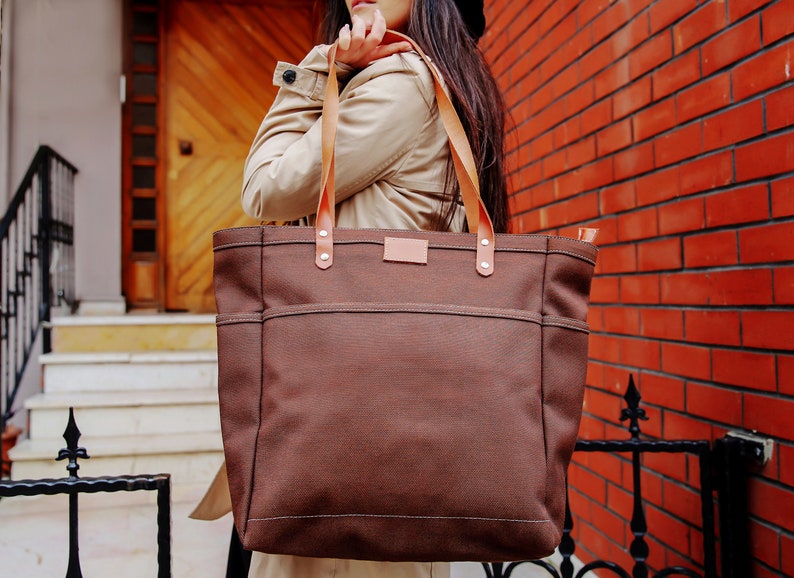 May include: A brown canvas tote bag with leather straps and a brown leather patch on the front. The bag is being held by a person.