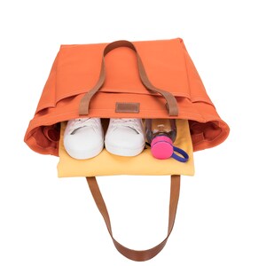 May include: Orange canvas tote bag with brown leather handles and a zippered pocket. The bag is open and contains two white sneakers, a clear water bottle with a pink lid, and a yellow cloth.