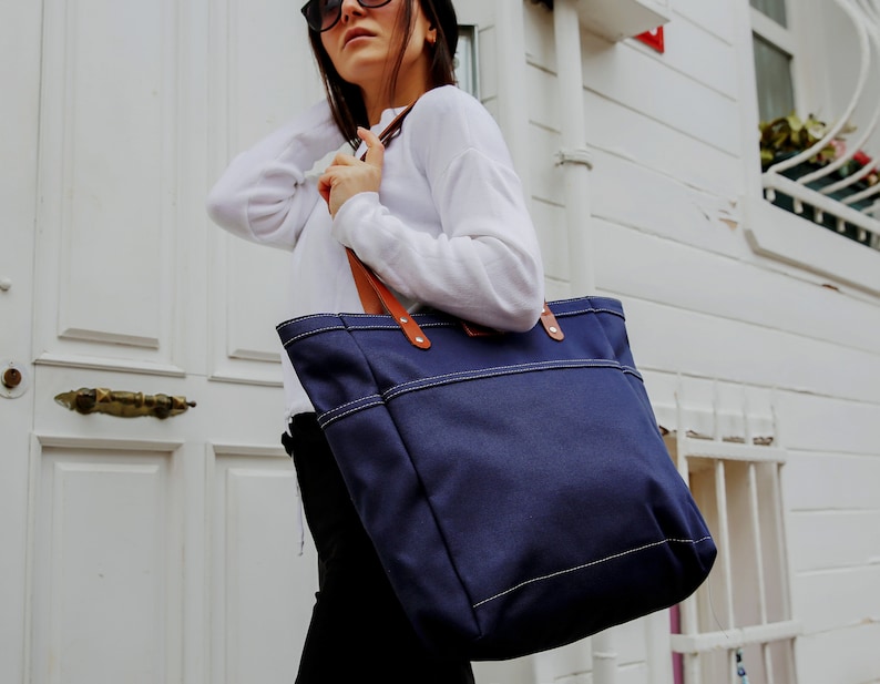 May include: A navy blue canvas tote bag with brown leather handles and white stitching. The bag is large and has a spacious interior.
