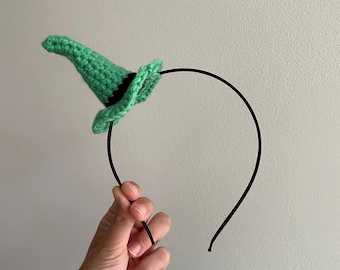 May include: A green crocheted witch hat on a black headband. The hat features a pointed shape and a black trim. The headband is thin and black. This handmade item is suitable for fancy dress or Halloween.