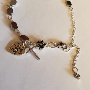 May include: A silver bracelet with a silver cross charm, a silver flower charm, and a silver heart charm that says "Made with Love". The bracelet is made of silver beads and a silver chain.