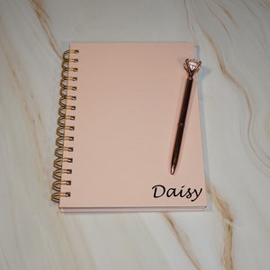 May include: A blush-colored spiral-bound notebook with a gold spiral and the name "Daisy" printed in black. A rose gold pen with a diamond-shaped top rests on the notebook. The notebook and pen are on a marble-patterned surface.
