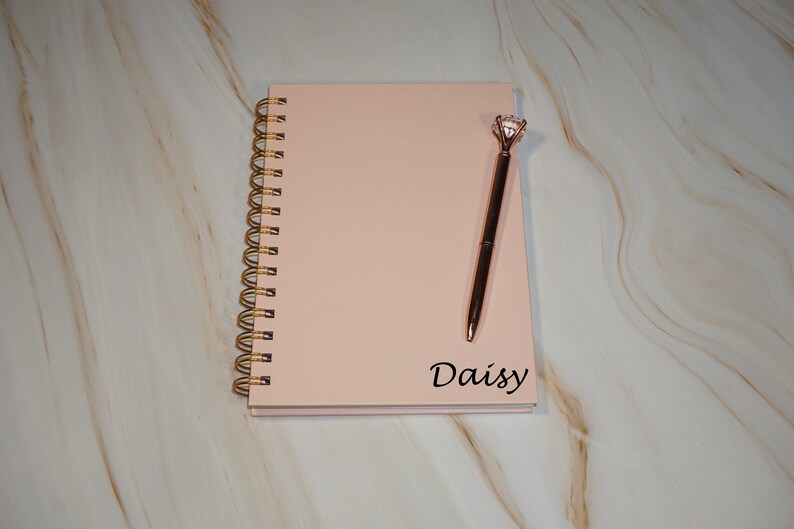 May include: A blush-colored spiral-bound notebook with a gold spiral and the name "Daisy" printed in black. A rose gold pen with a diamond-shaped top rests on the notebook. The notebook and pen are on a marble-patterned surface.
