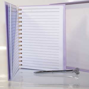 May include: A lavender spiral-bound notebook with lined pages, open to display the interior. A silver pen with a decorative top rests on the surface. The notebook is in a matching lavender box with a marble-patterned accent.
