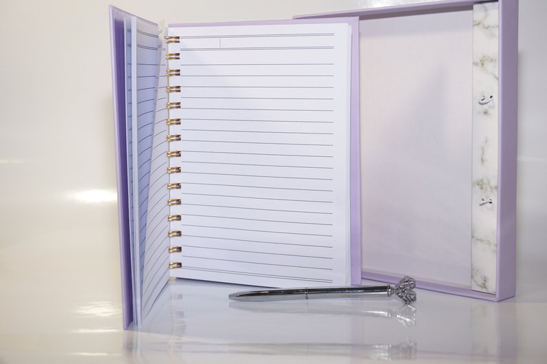May include: A lavender spiral-bound notebook with lined pages, open to display the interior. A silver pen with a decorative top rests on the surface. The notebook is in a matching lavender box with a marble-patterned accent.