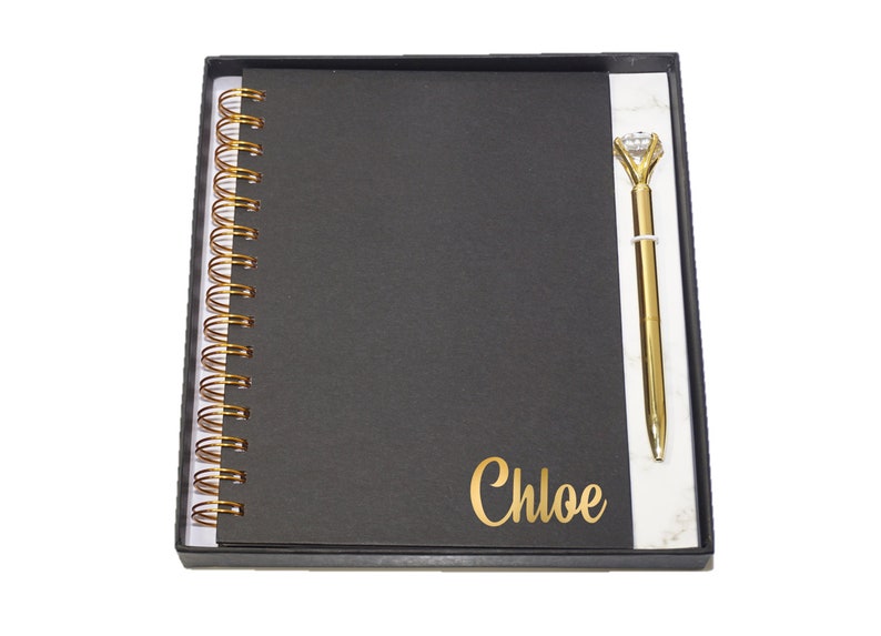 May include: A black spiral-bound notebook and a gold pen with a diamond-shaped top are presented in a black box. The notebook has the name "Chloe" in gold lettering. The pen is gold-colored.